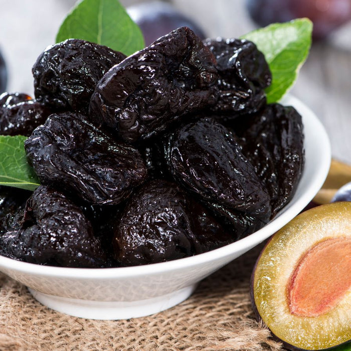 Ceramic bowl holds dark prunes beside fresh plum halves and green leaf.