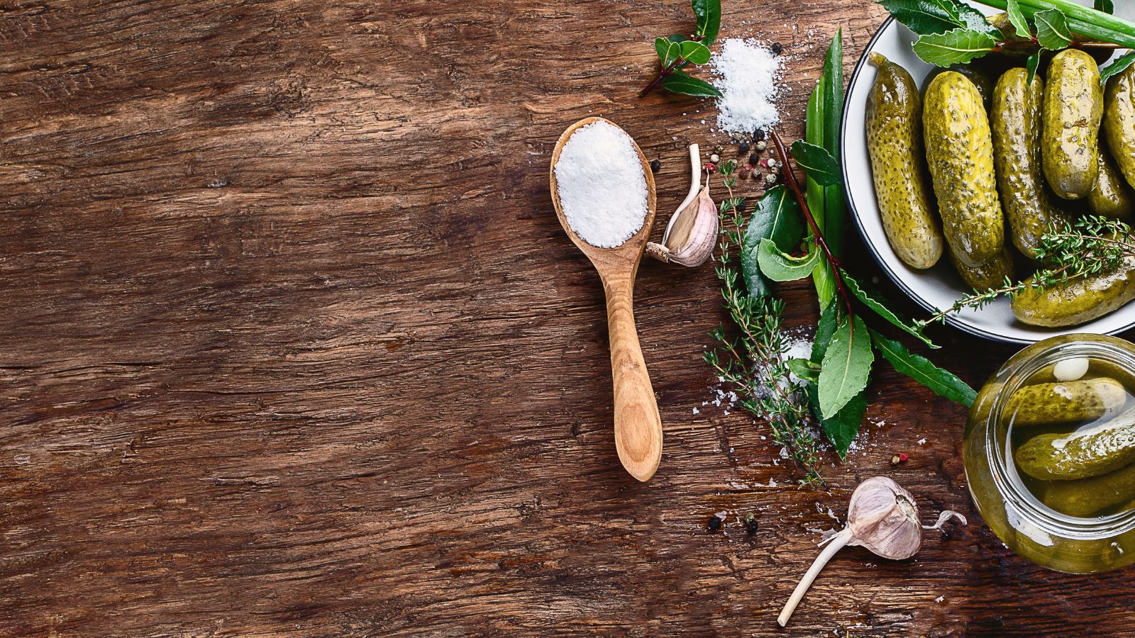 Wooden spoon of coarse salt beside pickles, garlic cloves and fresh herbs.