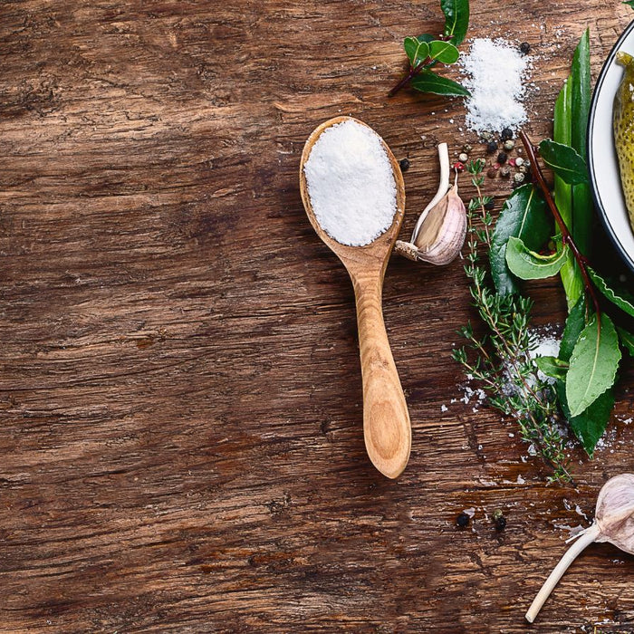 Wooden spoon of coarse salt beside pickles, garlic cloves and fresh herbs.