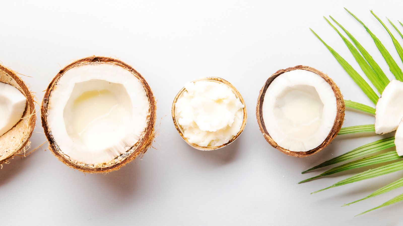 Halved coconuts with coconut oil and milk arranged on a white surface.