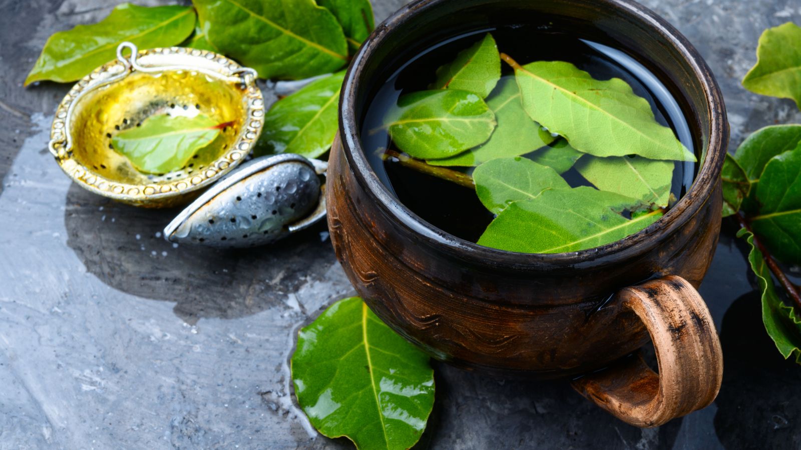Bay leaves steeping in herbal tea with metal infuser beside mug.