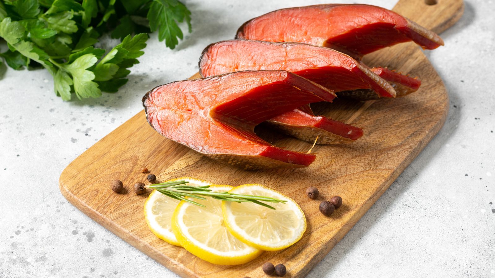 Sliced smoked salmon steaks on wooden board with lemon and herbs.