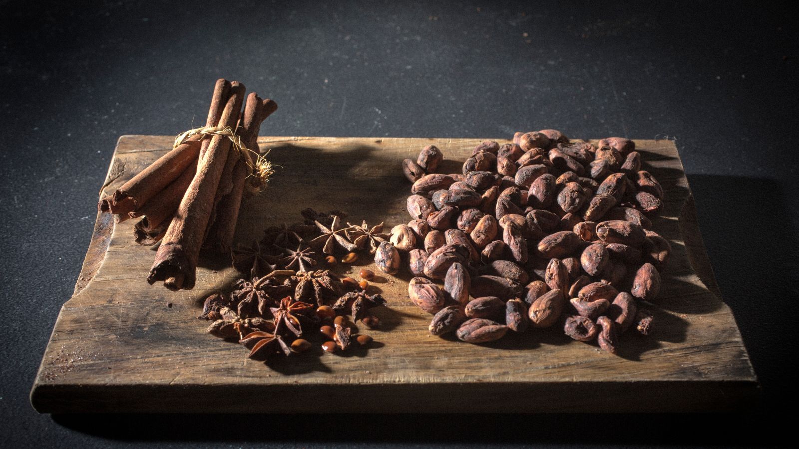 Cinnamon sticks and cacao beans on a rustic wooden board in dim light.