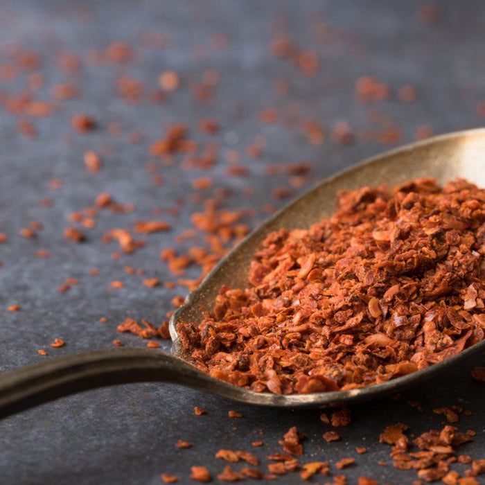 Dried crushed aleppo pepper on a metal spoon against dark surface.