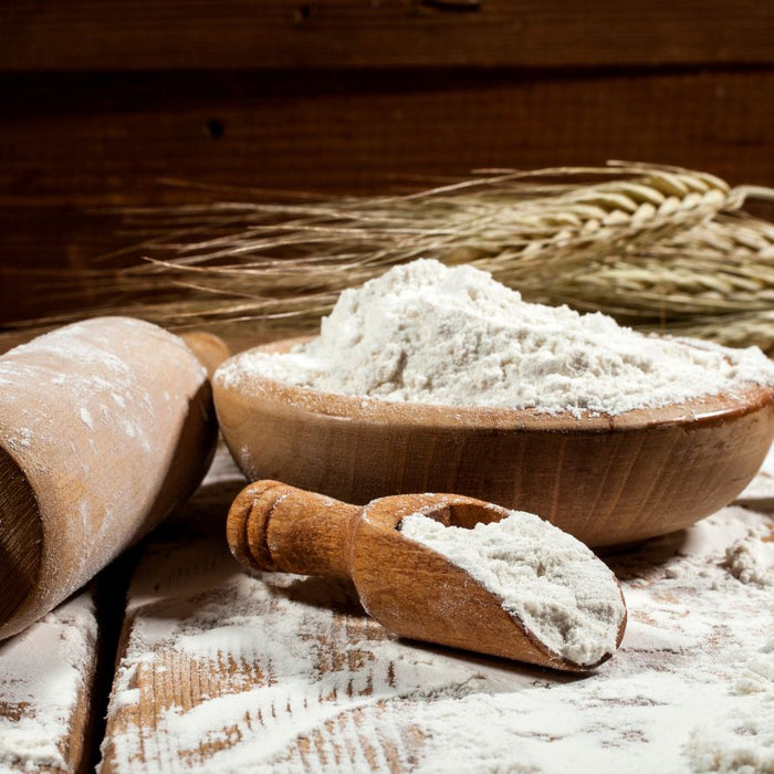 Wooden bowl of flour with rolling pin, wheat, and sifter on table.