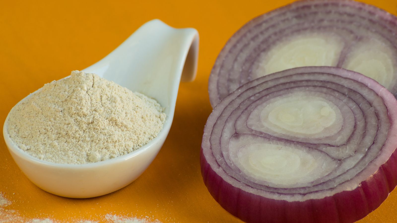Onion powder in spoon next to fresh red onion slices on yellow background.