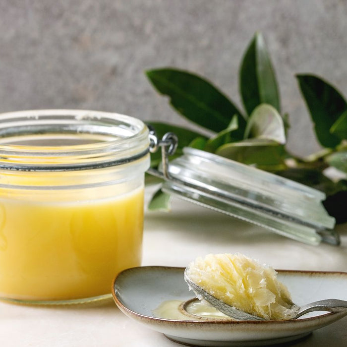 Glass jar of ghee with spoonful and leaves on white table.