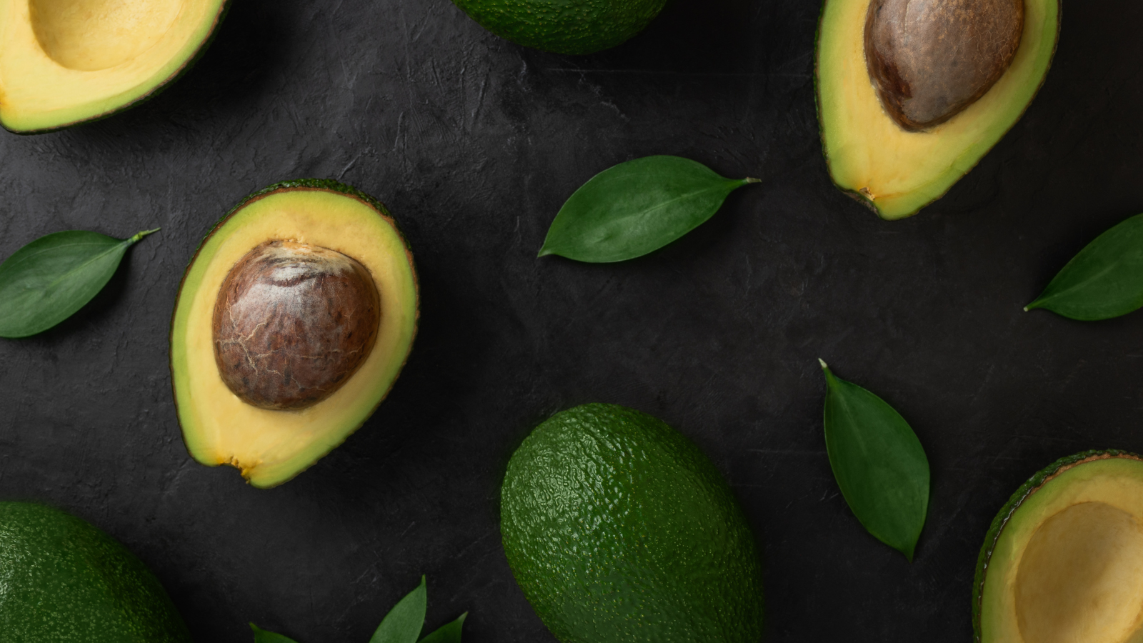Flat lay of halved and whole avocados with green leaves on dark table.