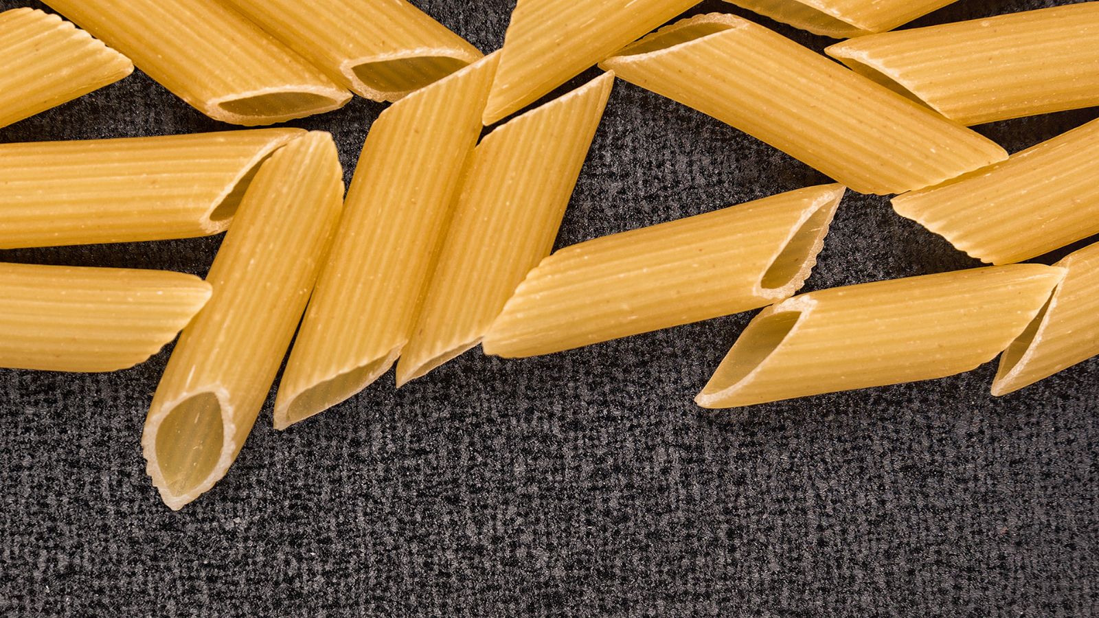 Close-up of uncooked penne pasta scattered on dark textured surface