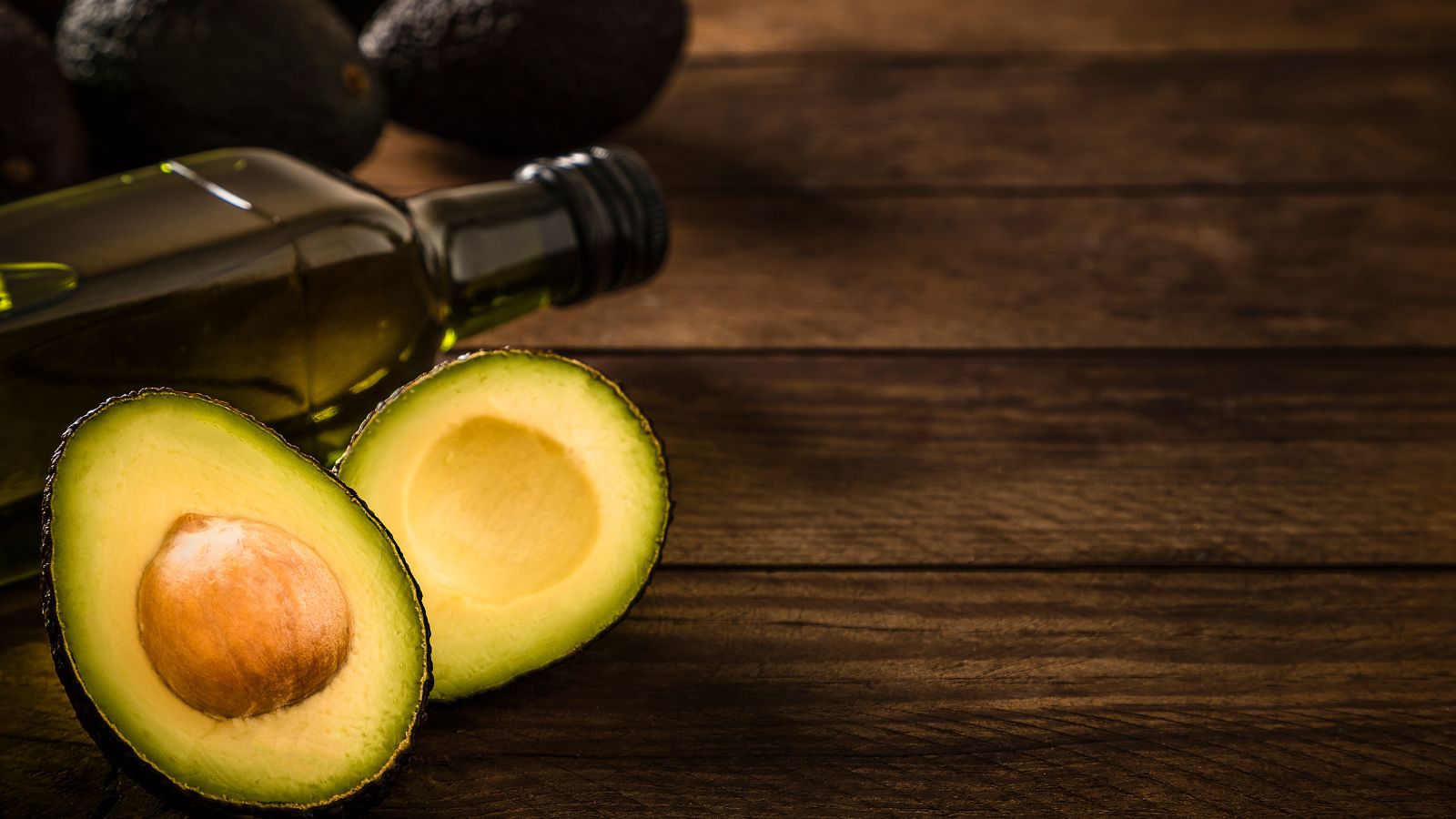 Avocado cut in half beside bottle of avocado oil on rustic wooden table