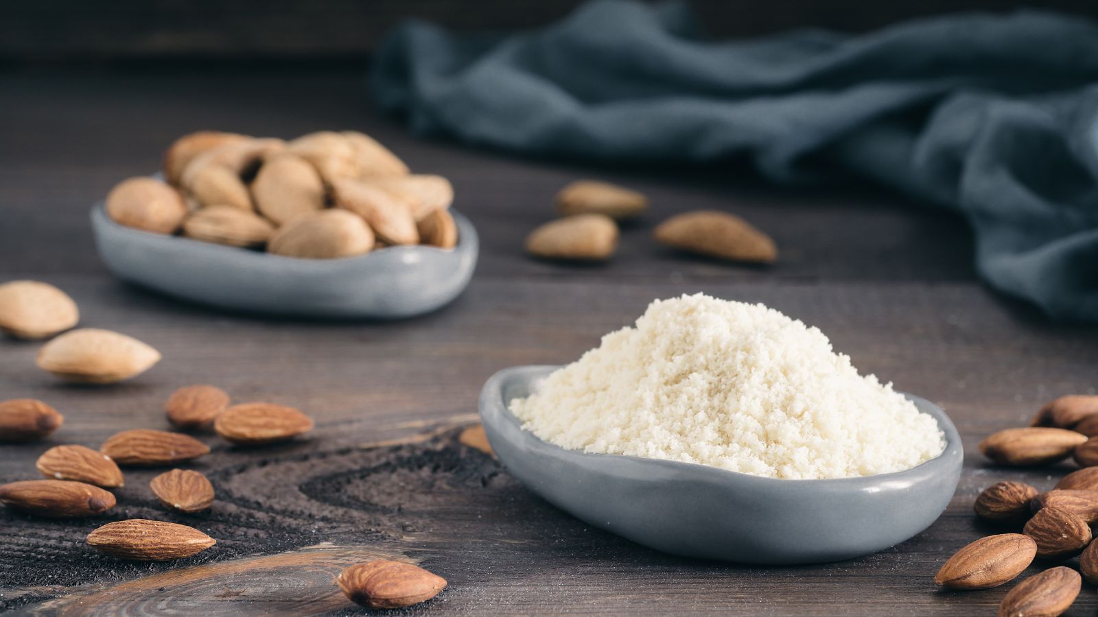 Bowl of almond flour powder beside scattered whole almonds on wood.