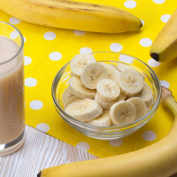 Glass of banana smoothie with straw next to bowl of slices and bananas.