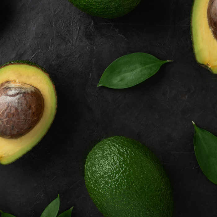 Flat lay of halved and whole avocados with green leaves on dark table.