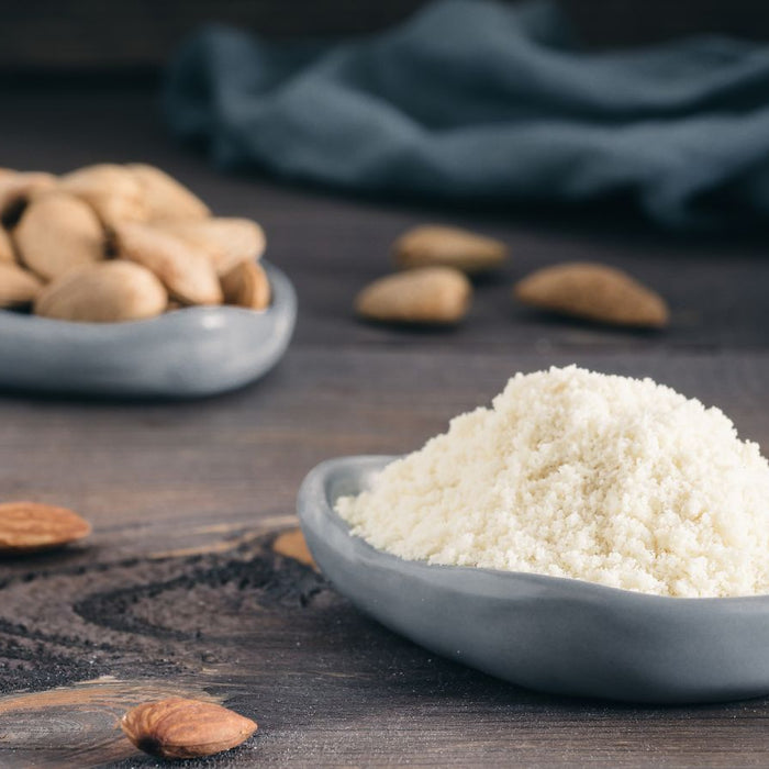 Bowl of almond flour powder beside scattered whole almonds on wood.