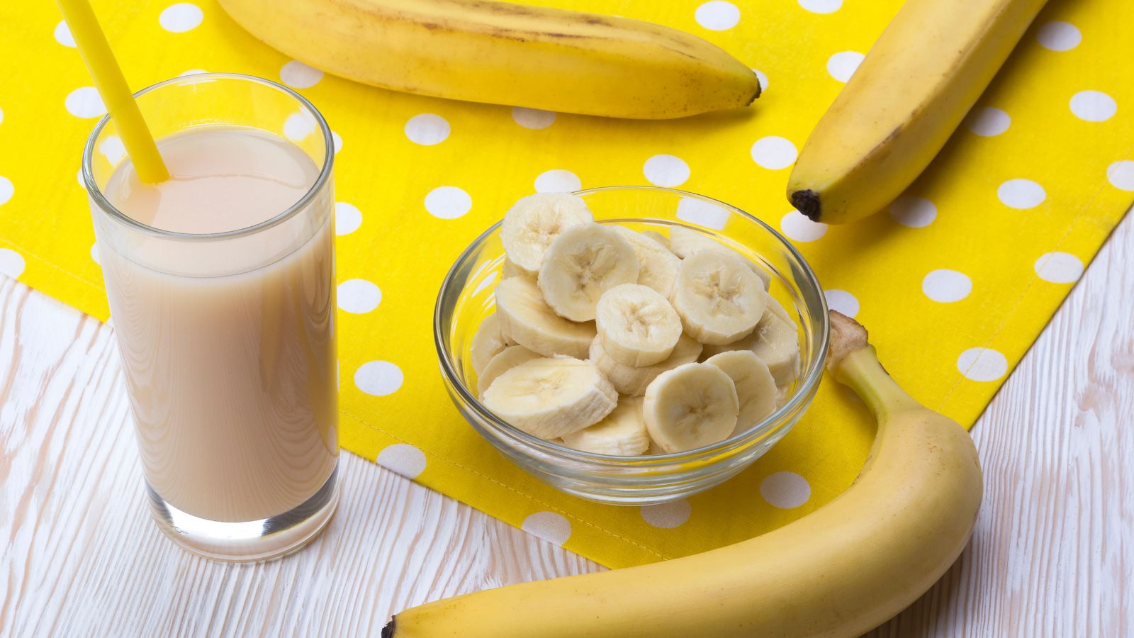 Glass of banana smoothie with straw next to bowl of slices and bananas.