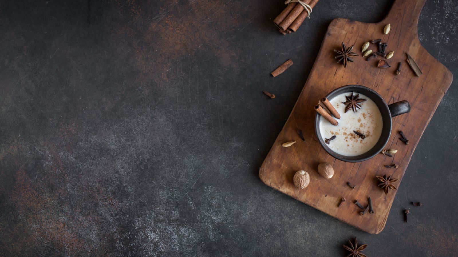 Warm chai tea in dark mug with star anise and spices on wooden board.