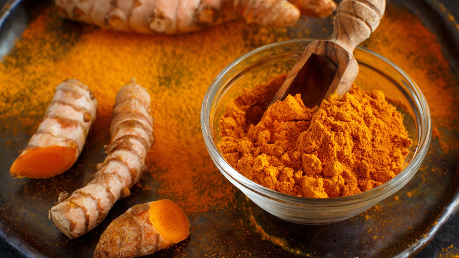 Bowl of turmeric powder with sliced roots on dark textured plate