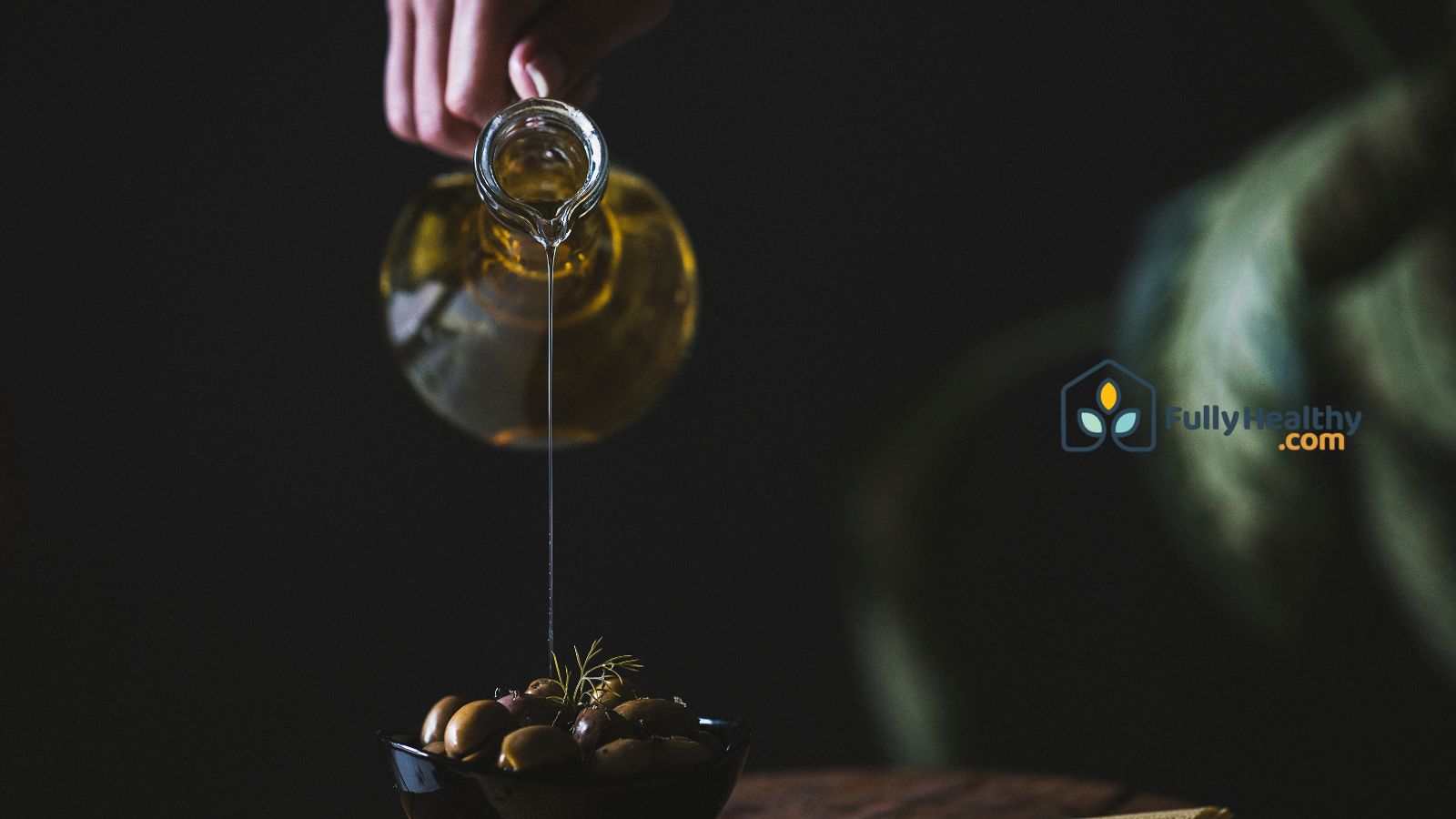 Pouring olive oil from glass jug over bowl of fresh ripe olives.