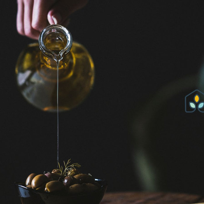 Pouring olive oil from glass jug over bowl of fresh ripe olives.