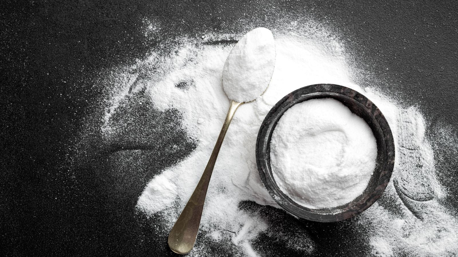 Spoon of baking soda beside spilled powder and bowl on dark surface.