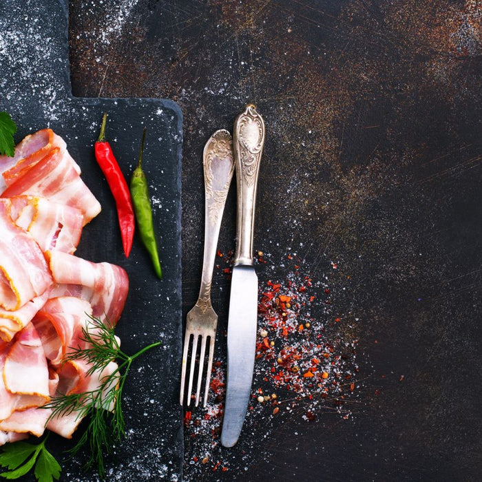 Raw bacon slices on cutting board with herbs and chili peppers.