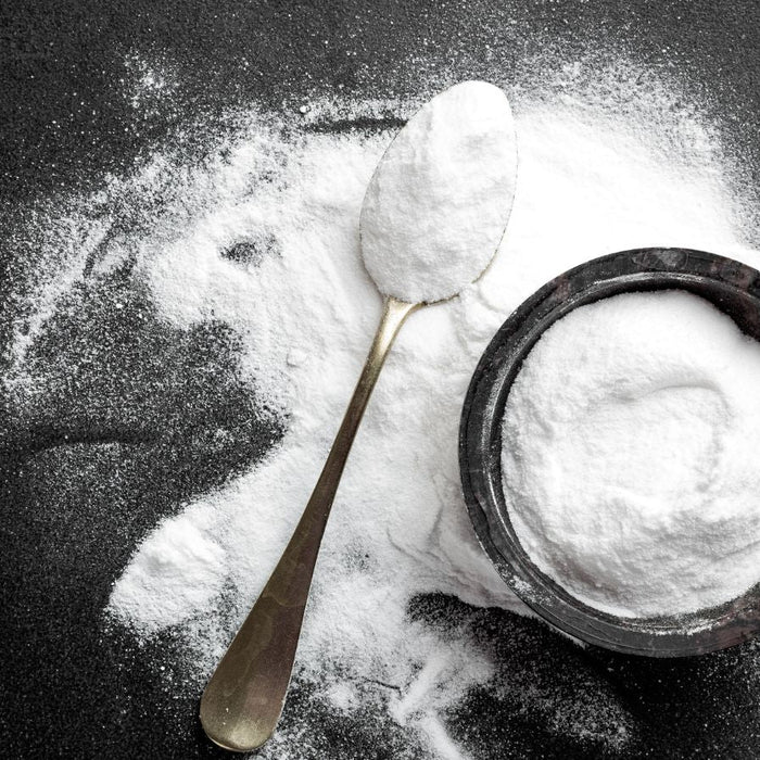 Spoon of baking soda beside spilled powder and bowl on dark surface.