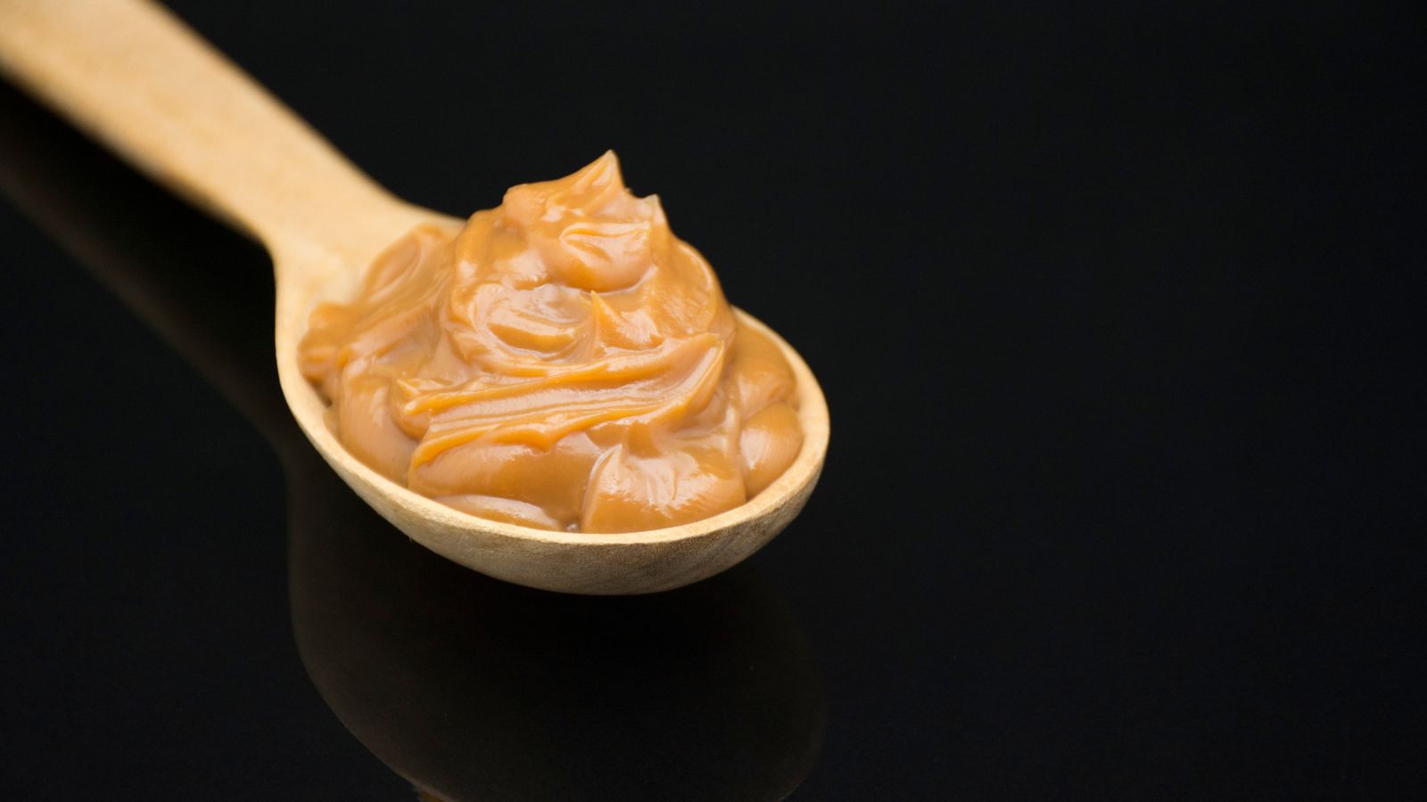 Wooden spoon with a swirl of smooth caramel on black background.