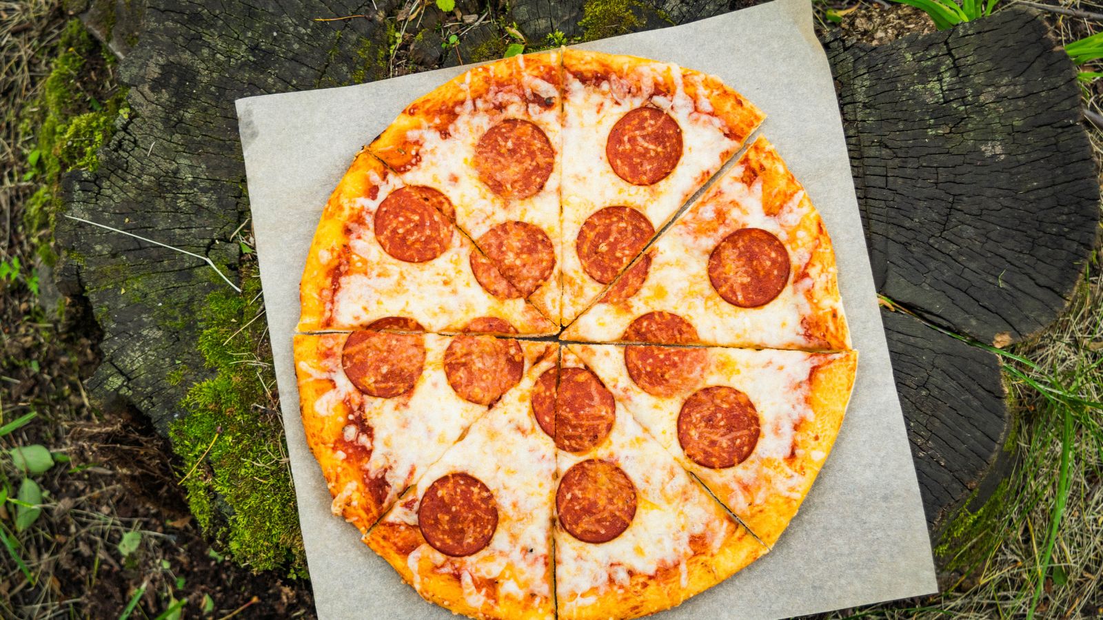 Pepperoni pizza on parchment paper atop tree stump outdoors.