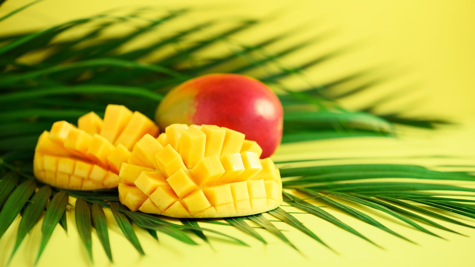 Two hedgehog-style mango halves and whole mango on palm leaf backdrop.