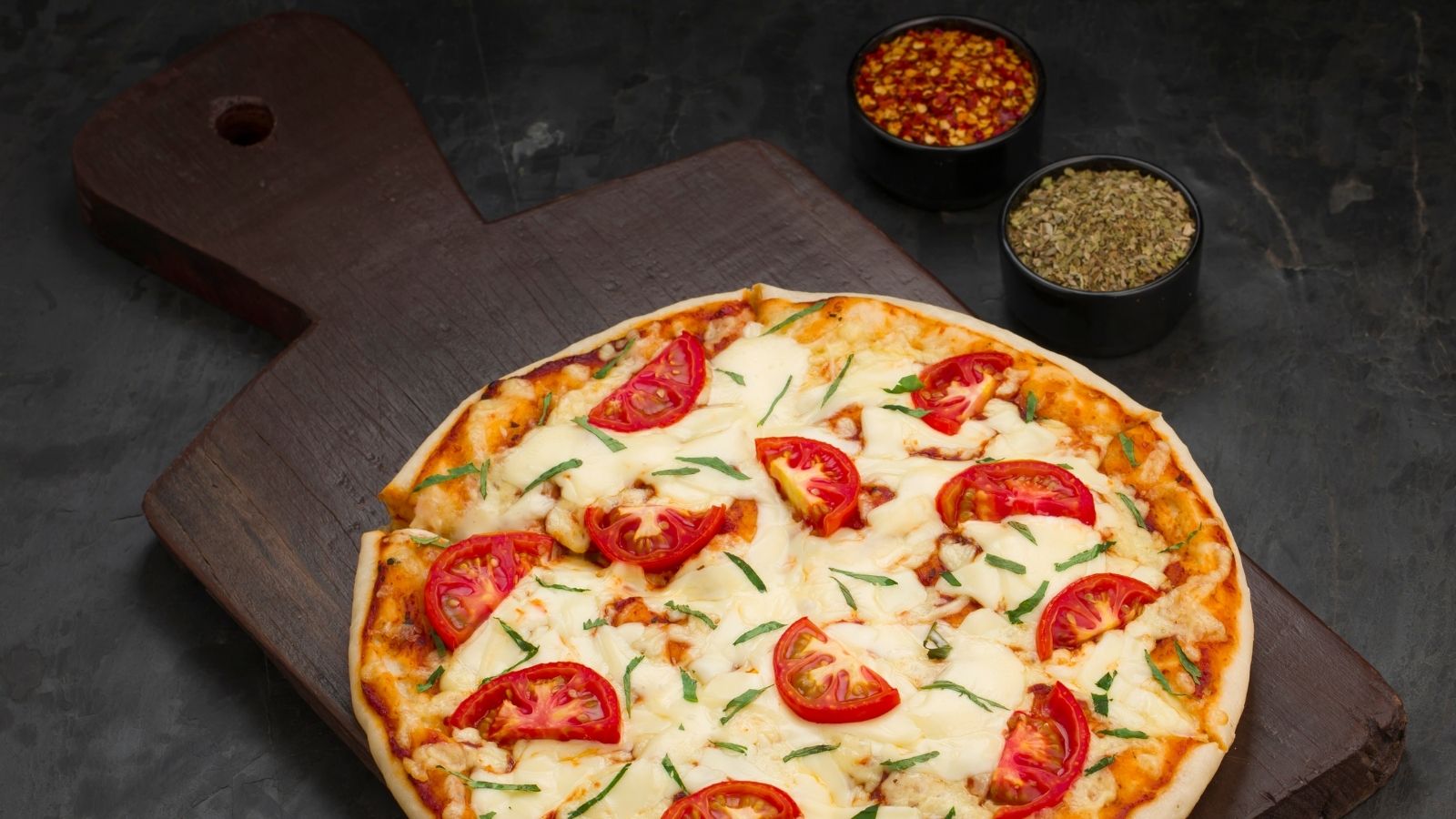 Tomato and cheese pizza on wooden board with herbs and spices bowls.