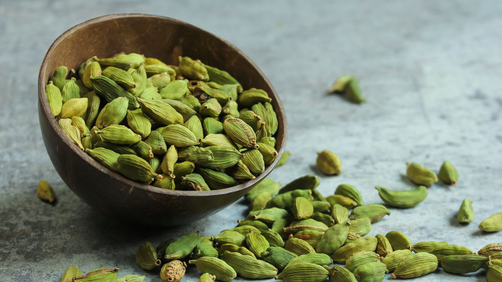 Fresh green cardamom pods in a wooden bowl on a gray surface.