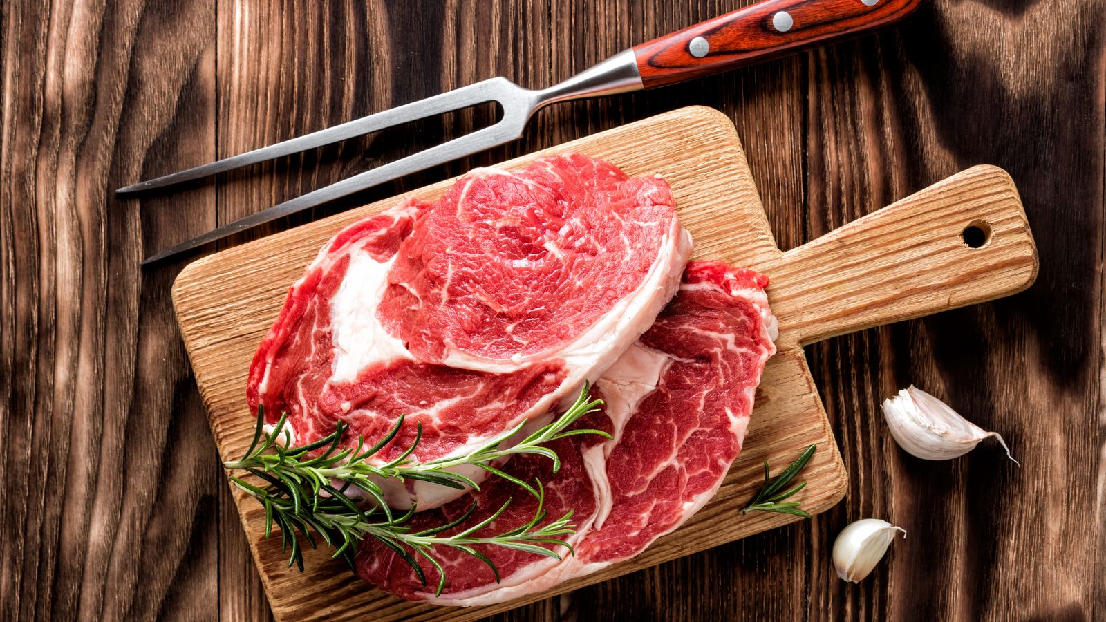 Raw ribeye steaks on wooden board with garlic and rosemary.