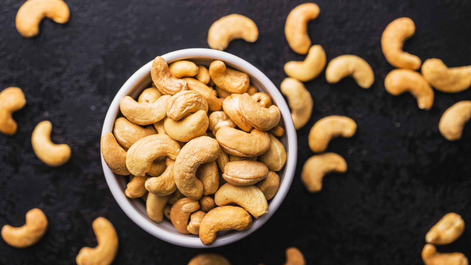Bowl of roasted cashew nuts on dark surface with scattered pieces.