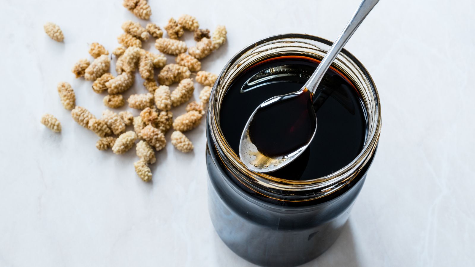Jar of dark molasses with a spoon and dried ingredients on a white surface.