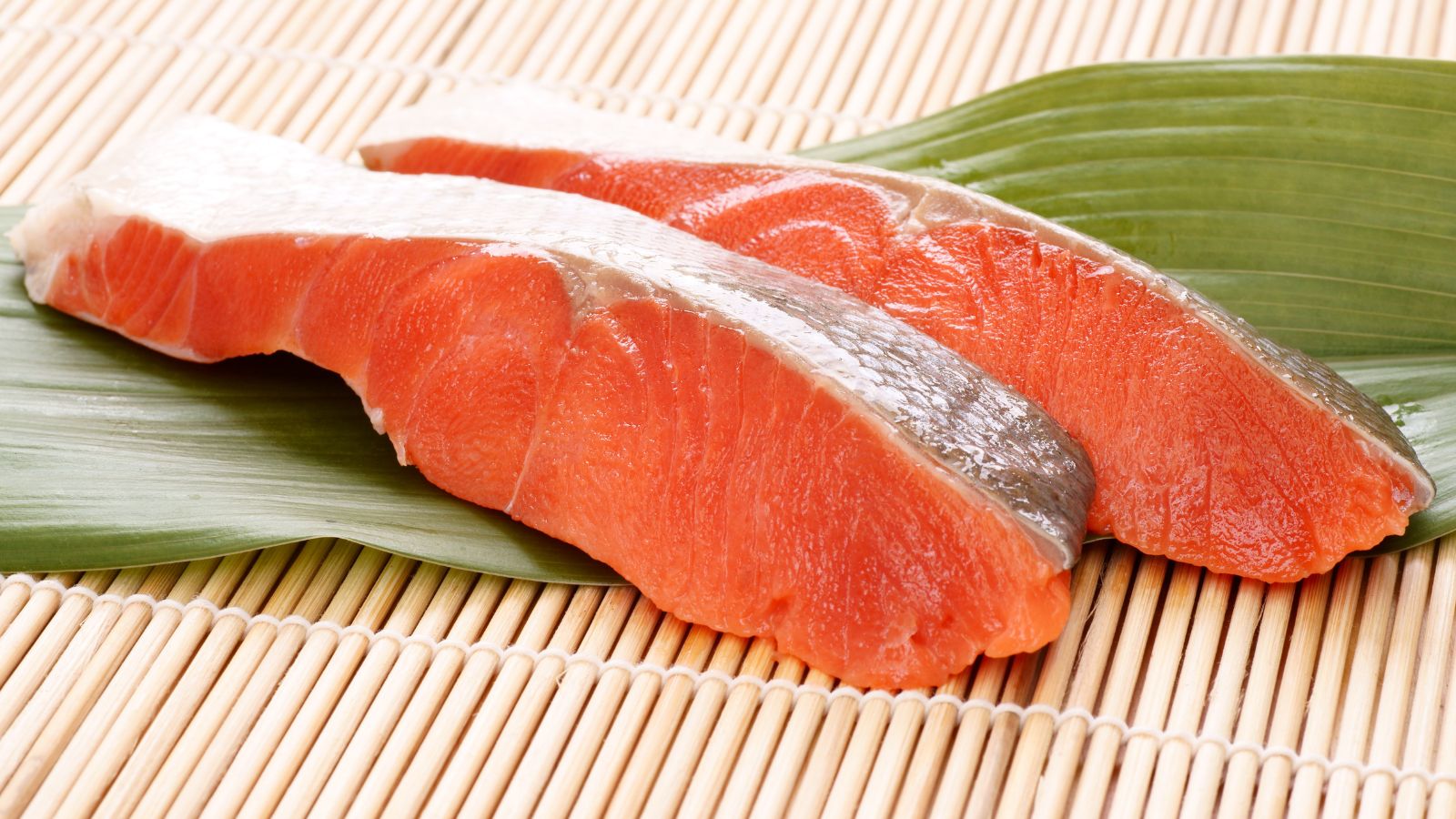 Fresh raw salmon fillets on bamboo mat and green leaf for sushi.