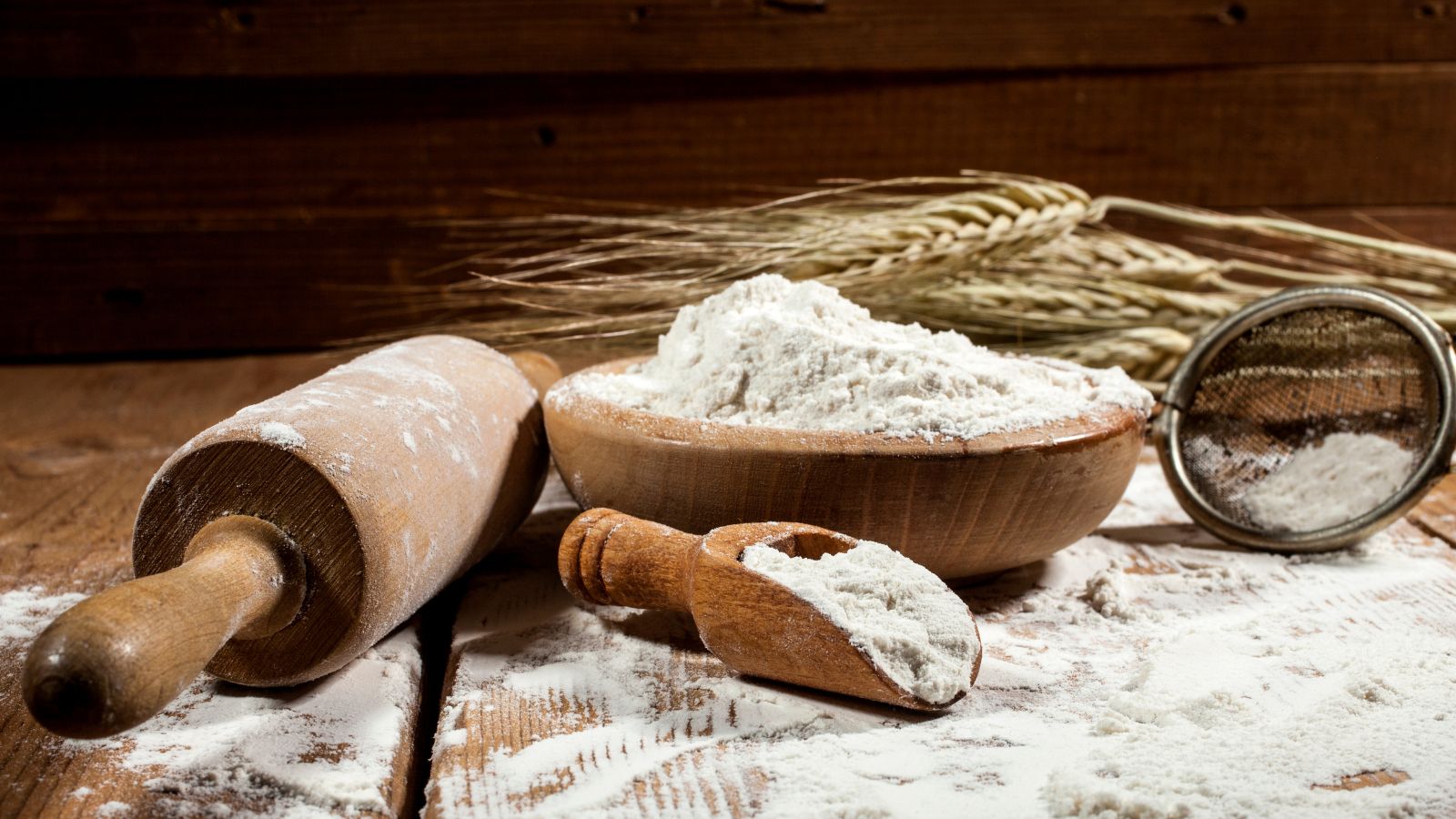 Wooden bowl of flour with rolling pin, wheat, and sifter on table.