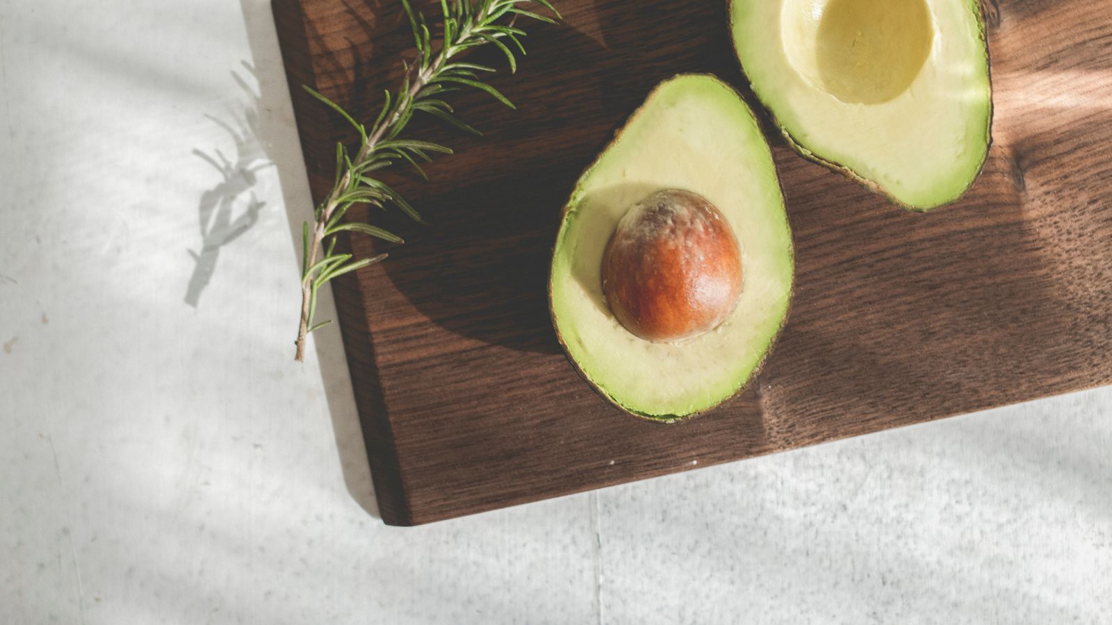 Avocado halves with seed and rosemary sprig on cutting board