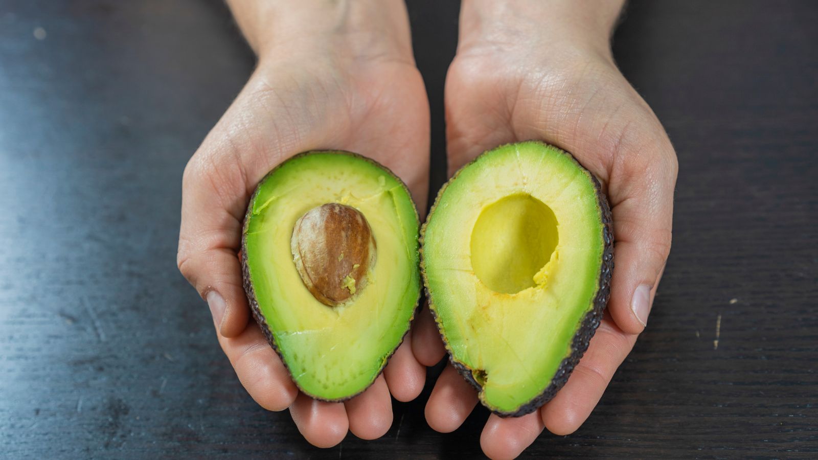 Hands holding avocado halves with seed on dark background