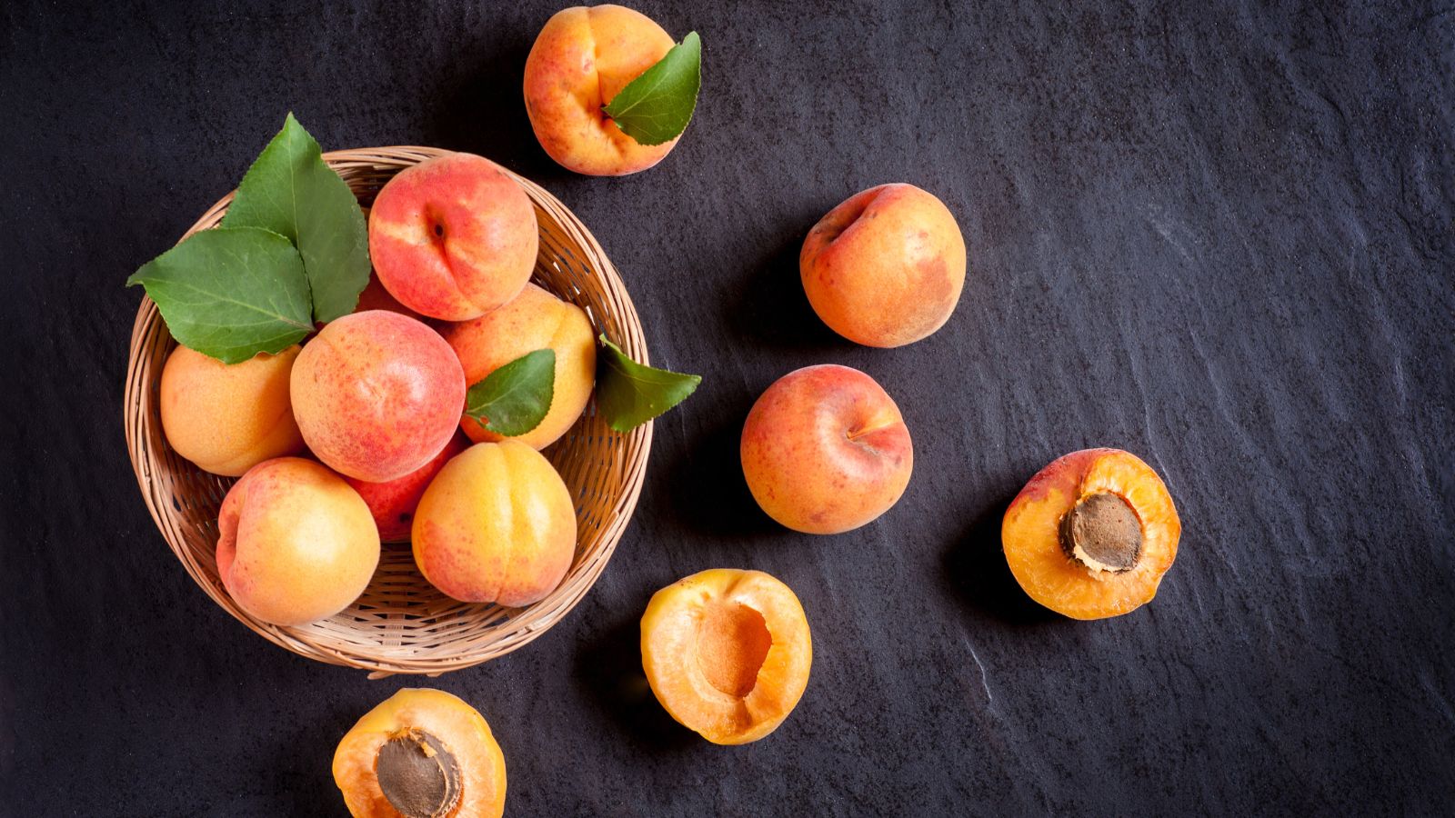 Basket of fresh apricots and halves on dark stone surface