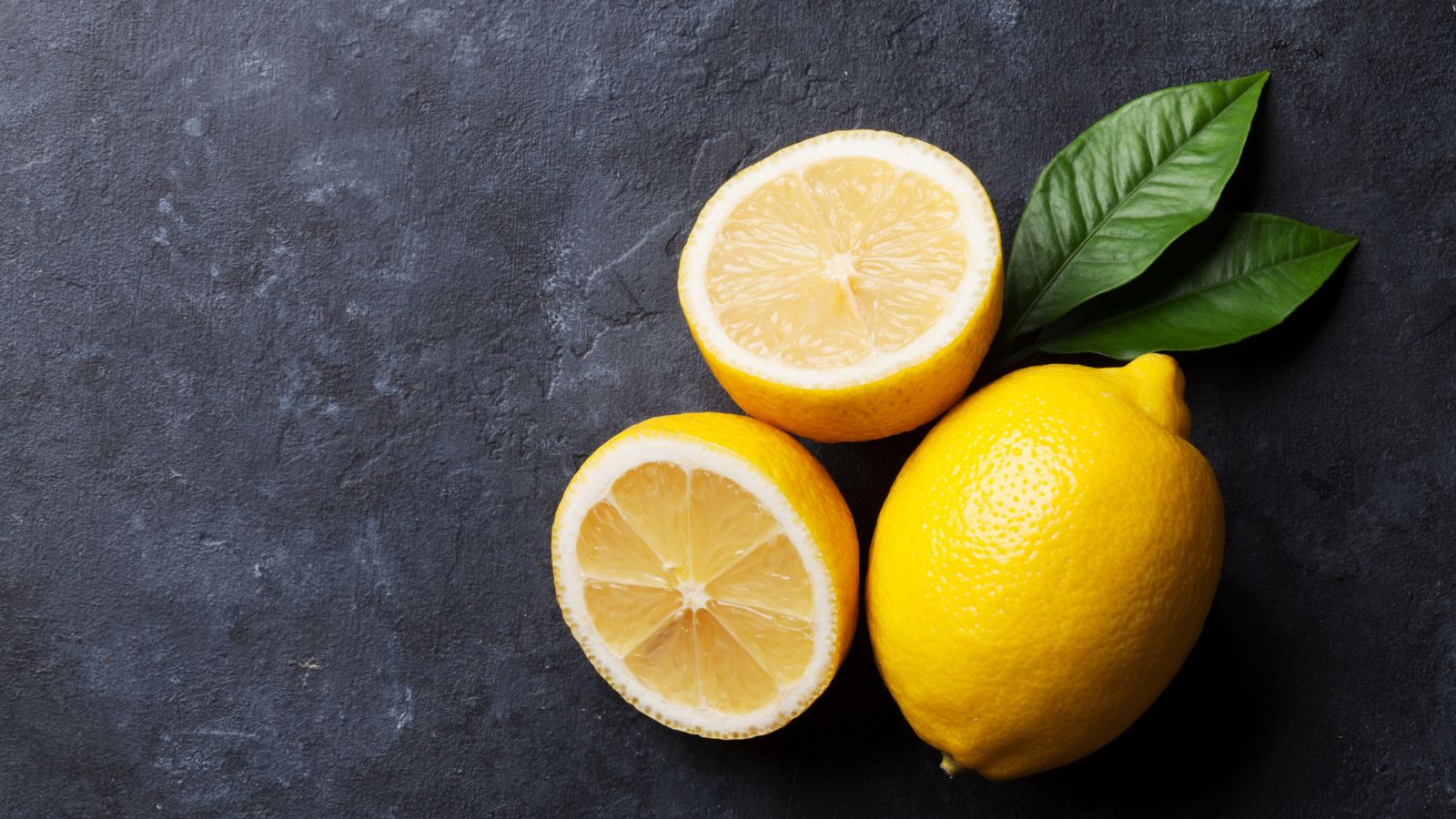 Whole and halved lemons with green leaves on dark surface