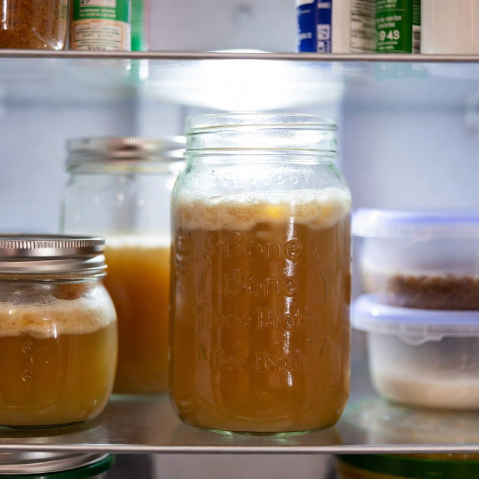 Jars of bone broth stored on a refrigerator shelf beside containers