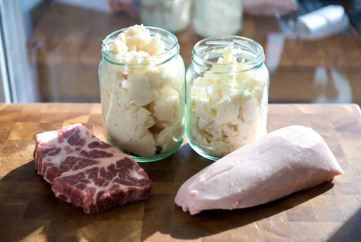 Jars of beef tallow beside raw beef and pork fat on wooden board