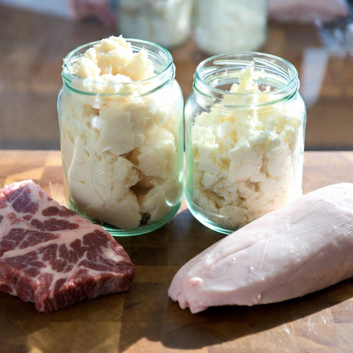 Jars of beef tallow beside raw beef and pork fat on wooden board