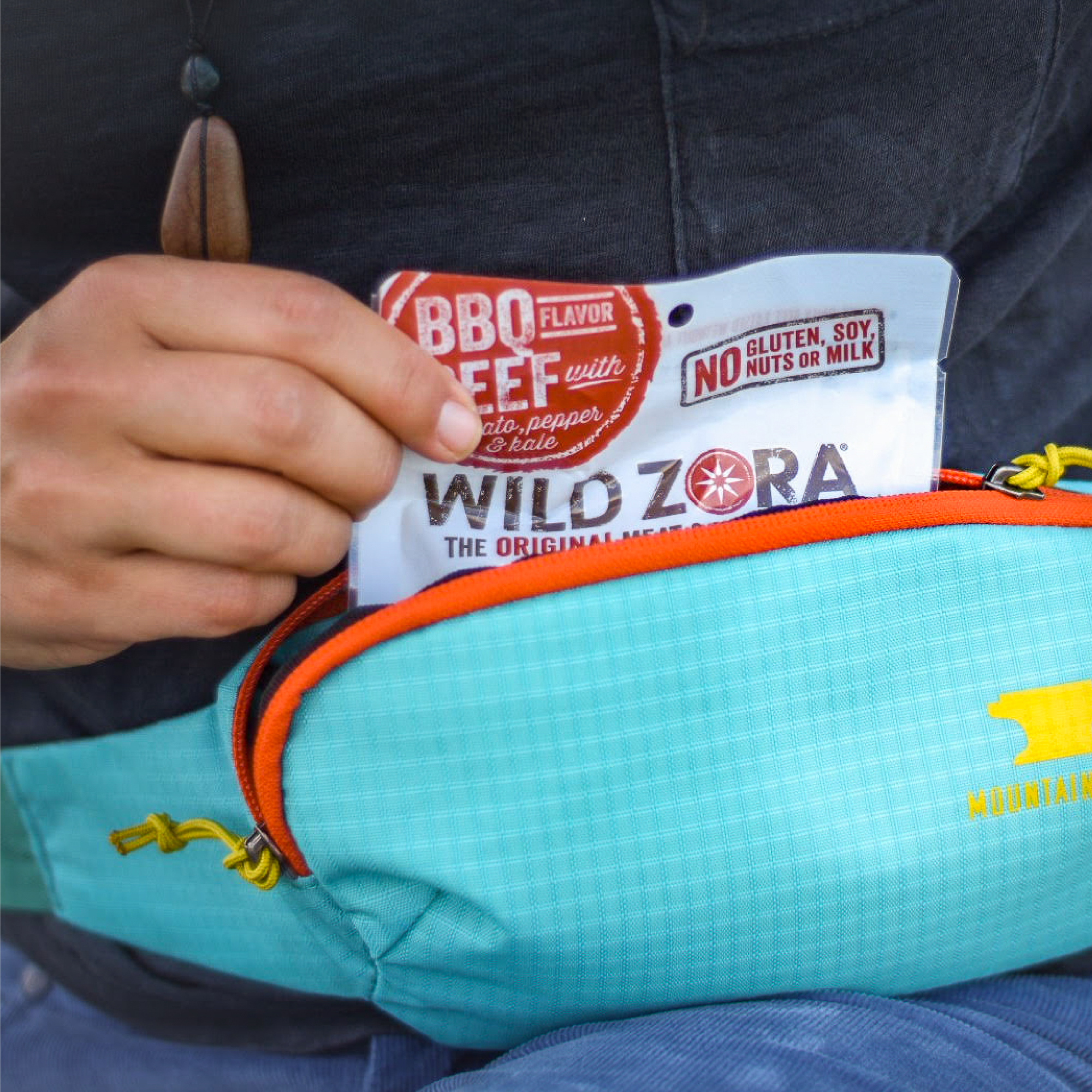 Person placing Wild Zora Meat & Veggie Bar BBQ Beef 1.1 oz into a blue waist pack for an on-the-go snack.